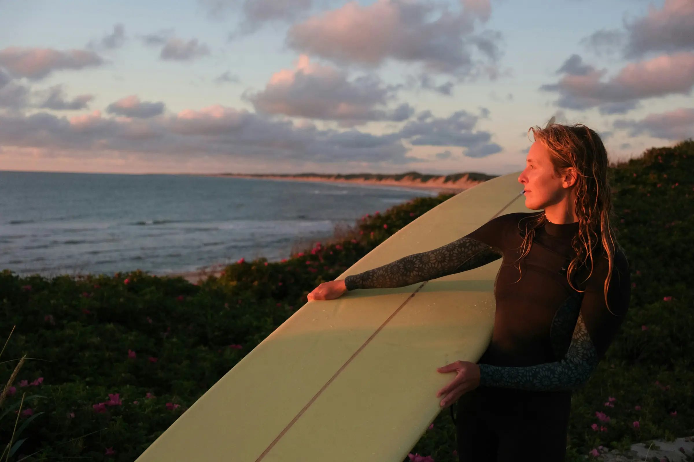 Ao fim do dia, uma surfista com a prancha debaixo do braço, a olhar o mar.
